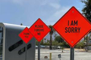Orange construction signs but with the messages "I AM SORRY", "PLEASE FORGIVE ME" and "THANK YOU" displayed on them.